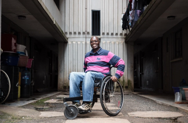 In a utility area below flats (apartments), a man smiles, with his hands on the push rims of his rough terrain wheelchair, ready to move.