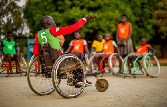 It’s a dry day on an outdoor basketball court, lined with trees. A male coach directs the team of your platers standing and seated in their sports wheelchairs.