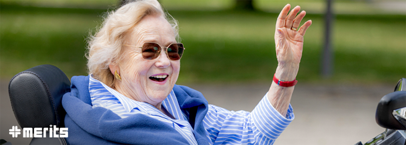 Older woman waving and smiling as she rides a power wheelchair outdoors